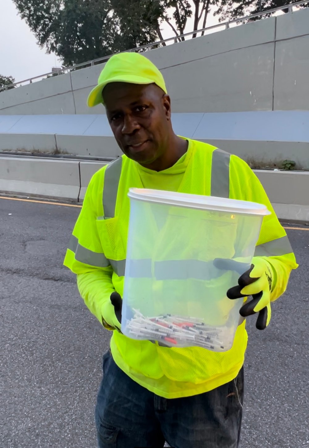At the Kingsbridge Underpass, a Black man wearing a neon green safety vest and protective gloves holds a case containing used syringes.